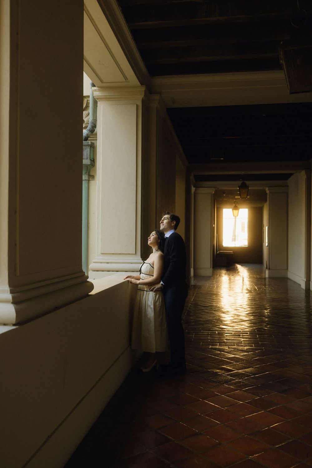 A couple stands in a sunlit corridor with terracotta floors and white columns.
