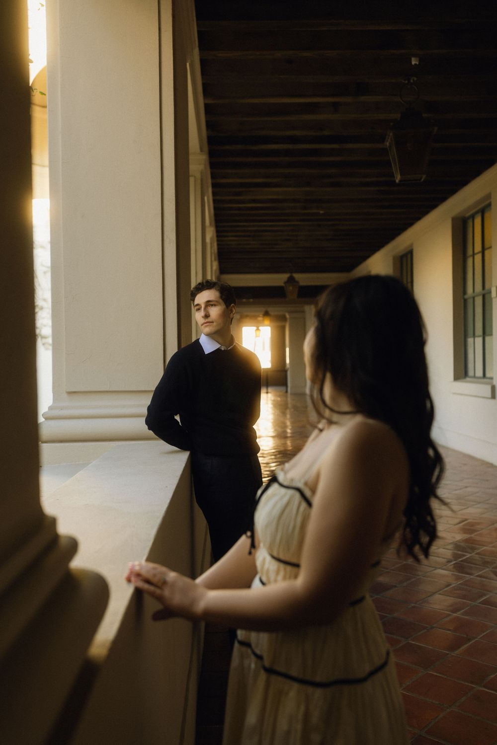 Two people stand in a sunlit corridor with Spanish colonial architecture featuring terra cotta floors and exposed wooden beams.