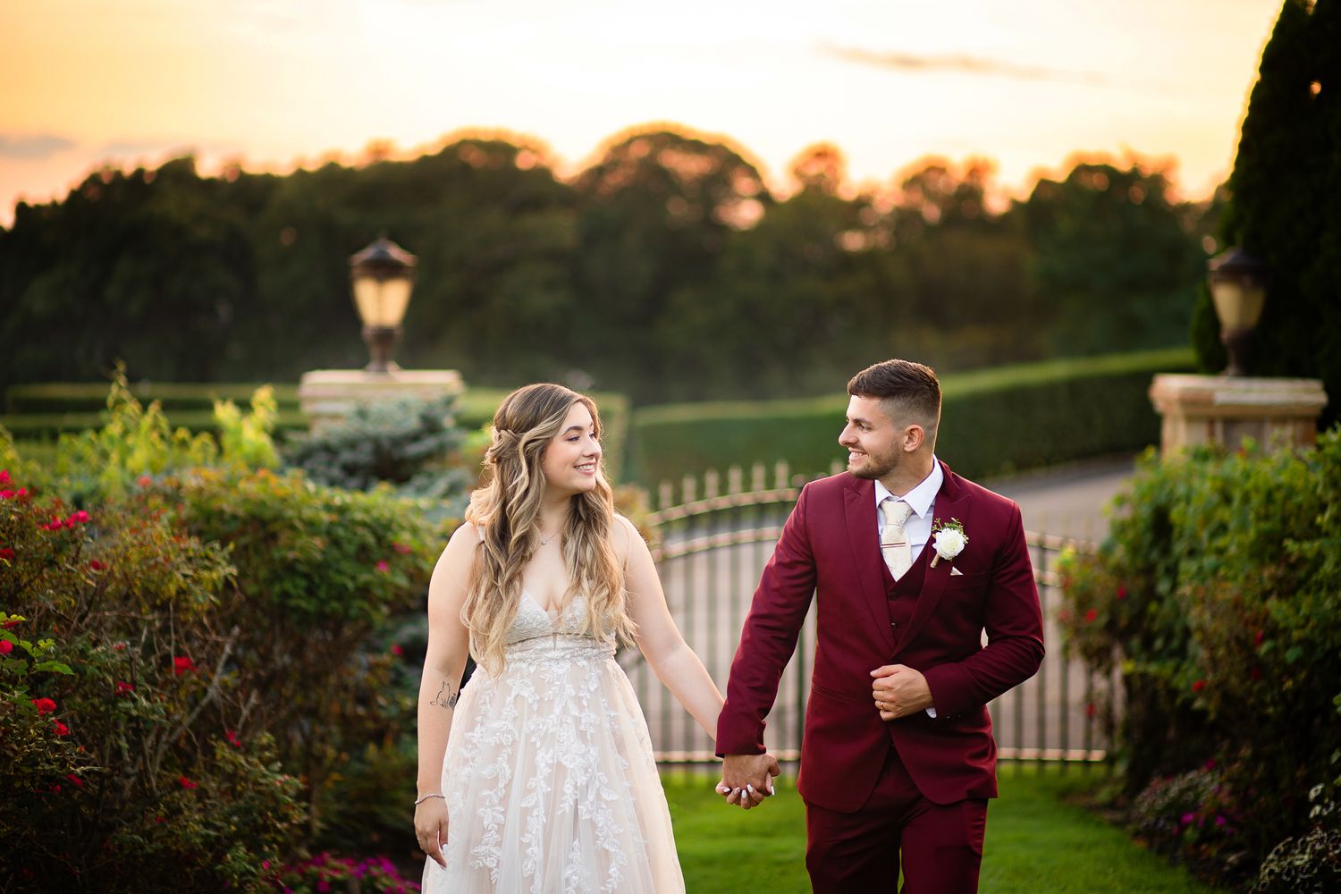 A couple in wedding attire walks hand in hand in a garden at sunset with a burgundy suit and white lace dress.