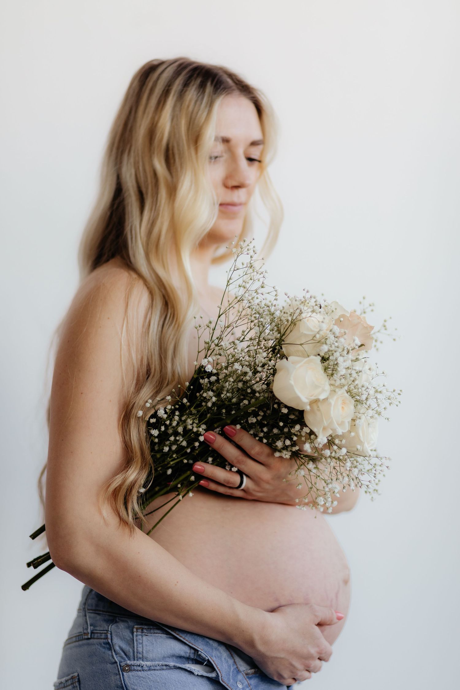 A pregnant woman in jeans holds a delicate bouquet of white roses and baby's breath against a light background.