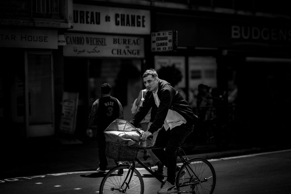 A black and white street photo of a cyclist riding past urban storefronts.