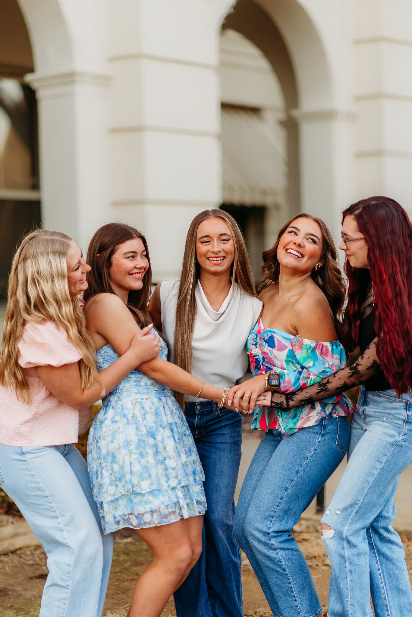 A group of friends laugh and embrace while wearing casual summer outfits against a stone archway background.