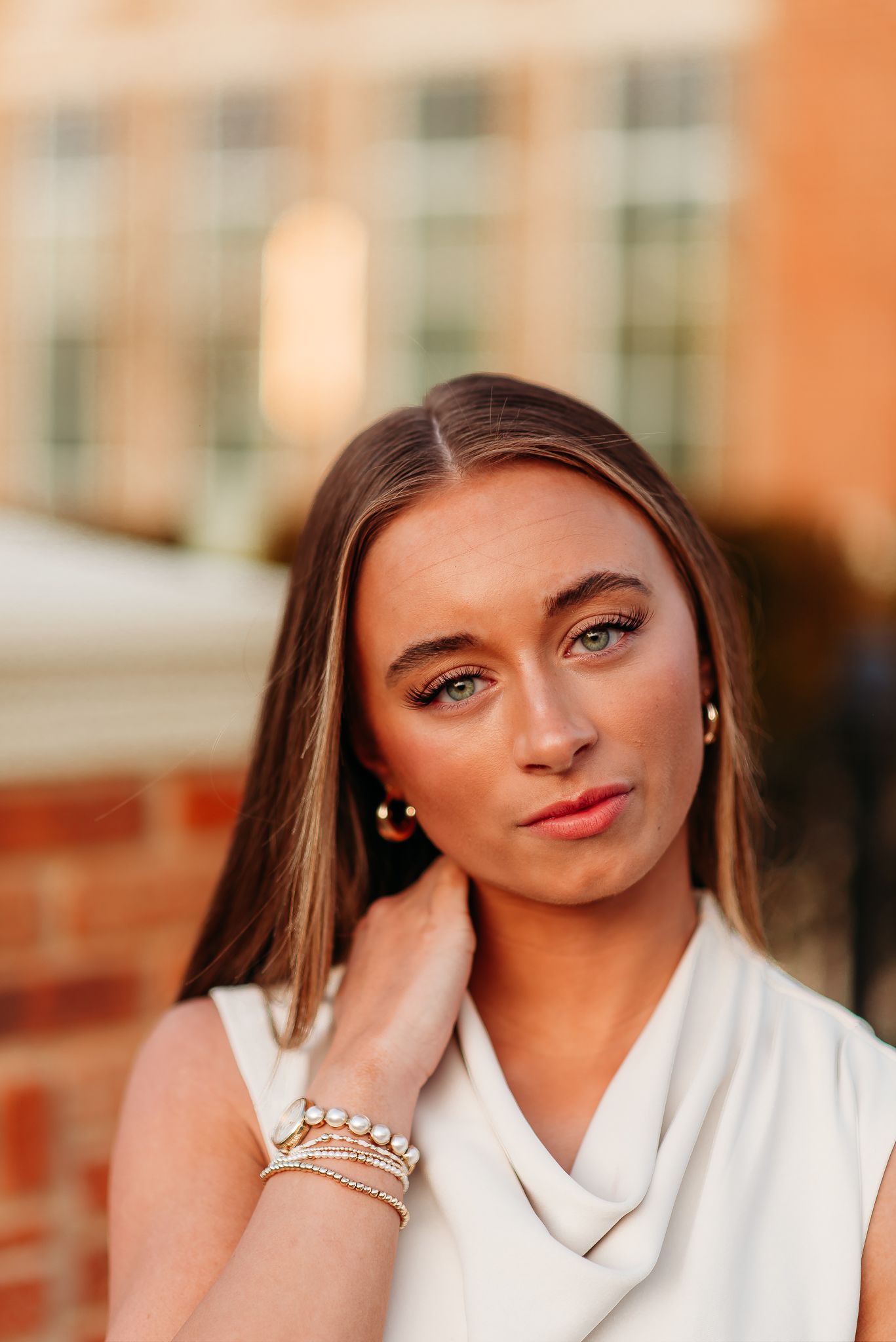 A close-up portrait with delicate jewelry and a white cowl neck top against brick architecture.
