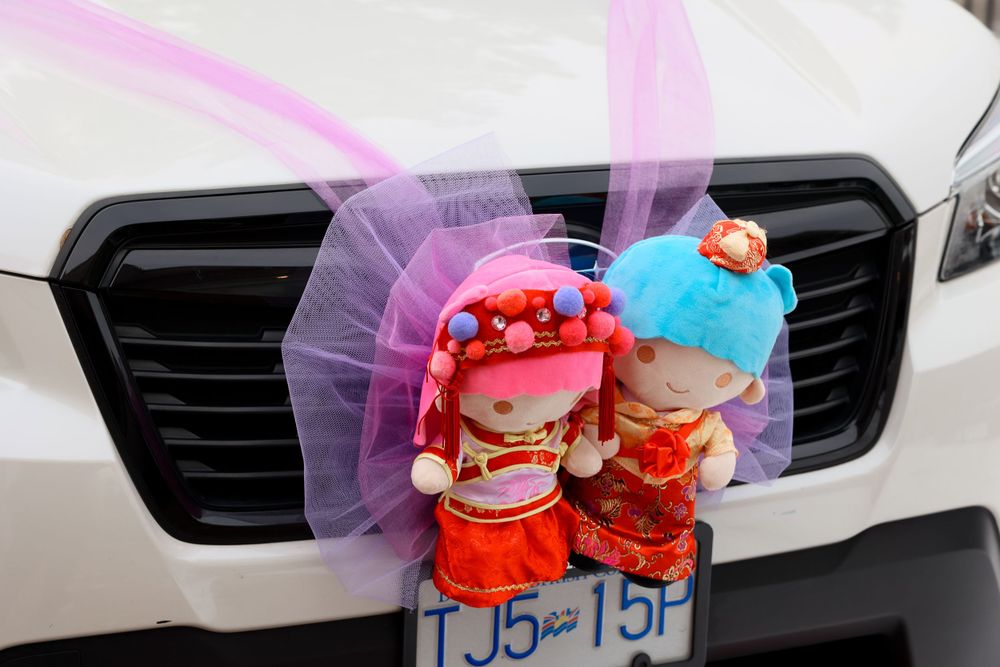 Two decorative dolls with colorful tulle attached to a car's front grille near the license plate.