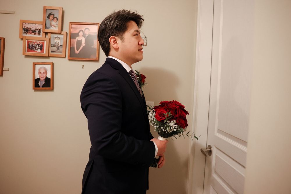 A person in a tuxedo holds a red bouquet while standing in a room with framed photos on the wall. Chinese wedding