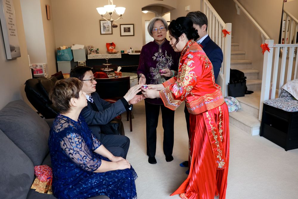 People gathered in a living room interact with someone wearing a traditional bright red Chinese dress during a celebration.