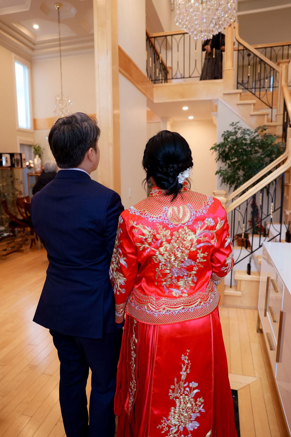 A couple in formal attire stand facing staircase, with person wearing ornate red traditional Chinese wedding dress with gold embroidery.