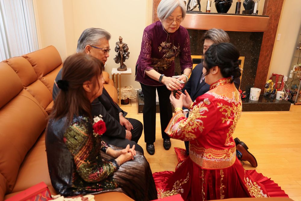 People gather in a living room during a traditional chinese tea ceremony wearing festive sweaters near a fireplace.