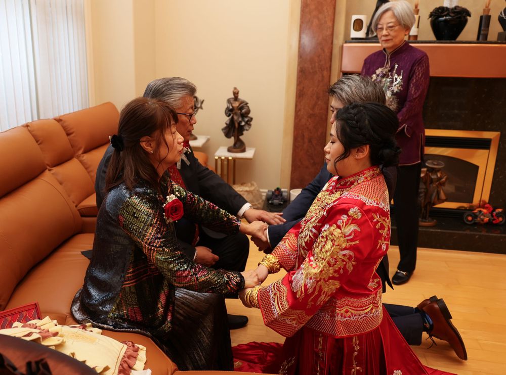 Three women in colorful traditional Asian dresses interact warmly in a living room setting with couch and fireplace.