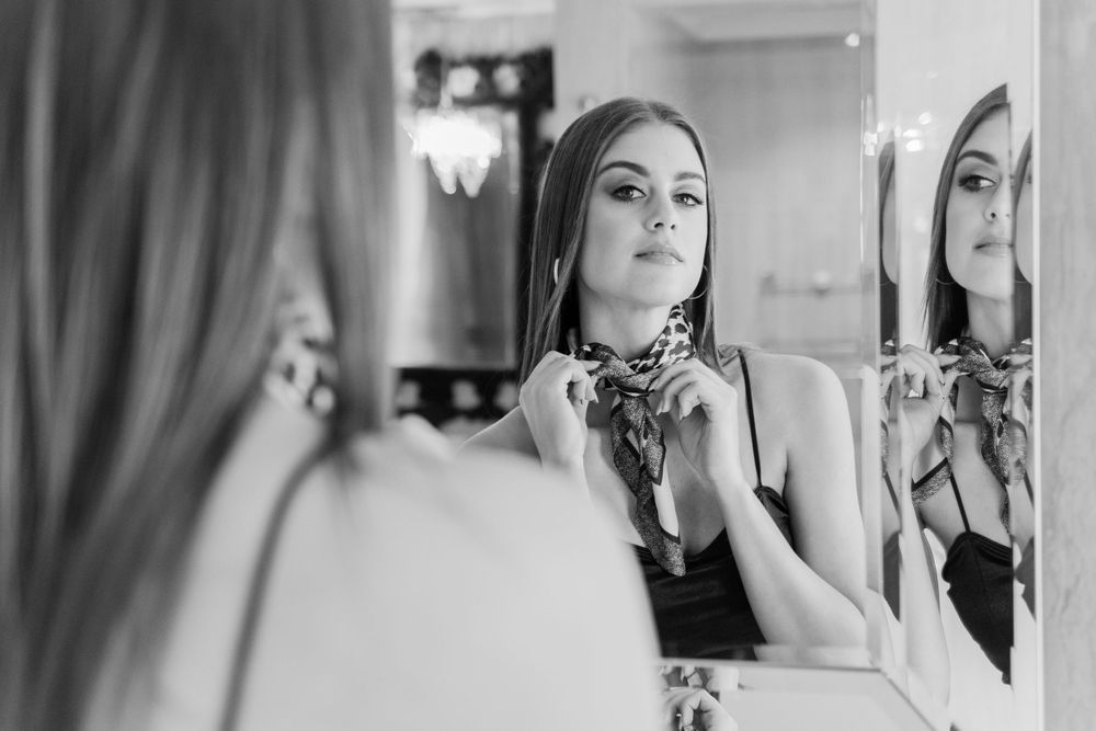 Black and white photo of a woman adjusting a scarf while looking at her reflection in a mirror.