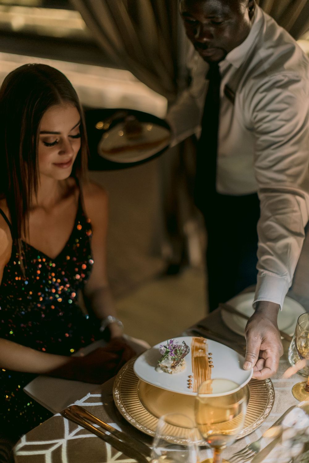 Waiter in white shirt serving elegant gold cutlery on white plate to woman in sequin dress at fine dining table.