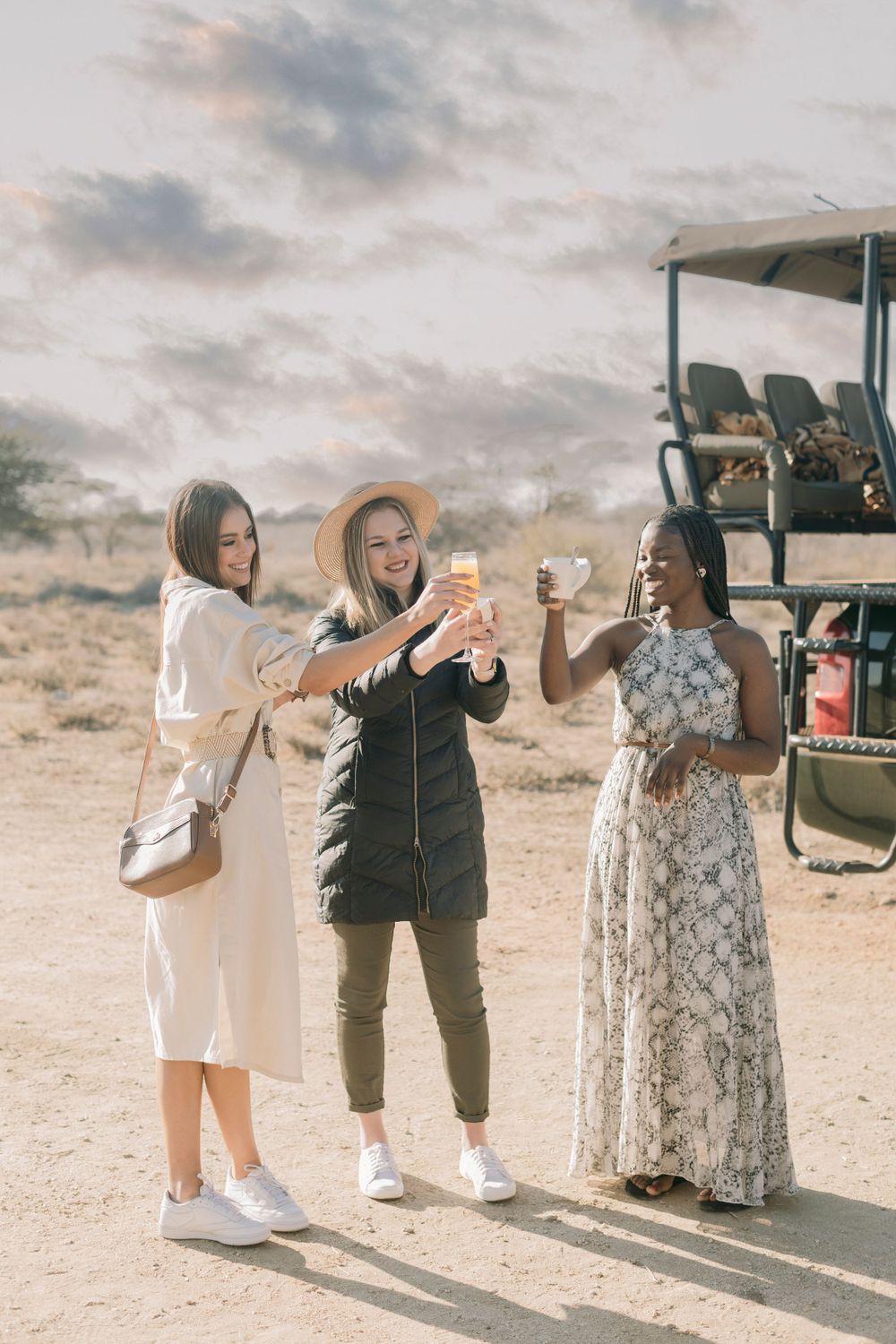 Three women toasting with drinks on an African safari, standing near a game drive vehicle in arid landscape.