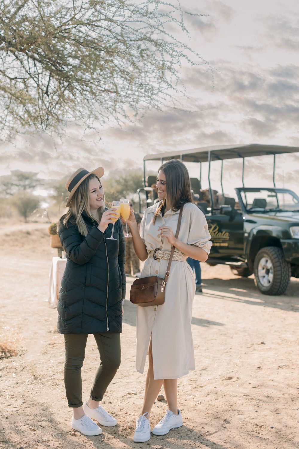 Two women enjoying drinks and laughing on an African safari with a game drive vehicle parked behind them.