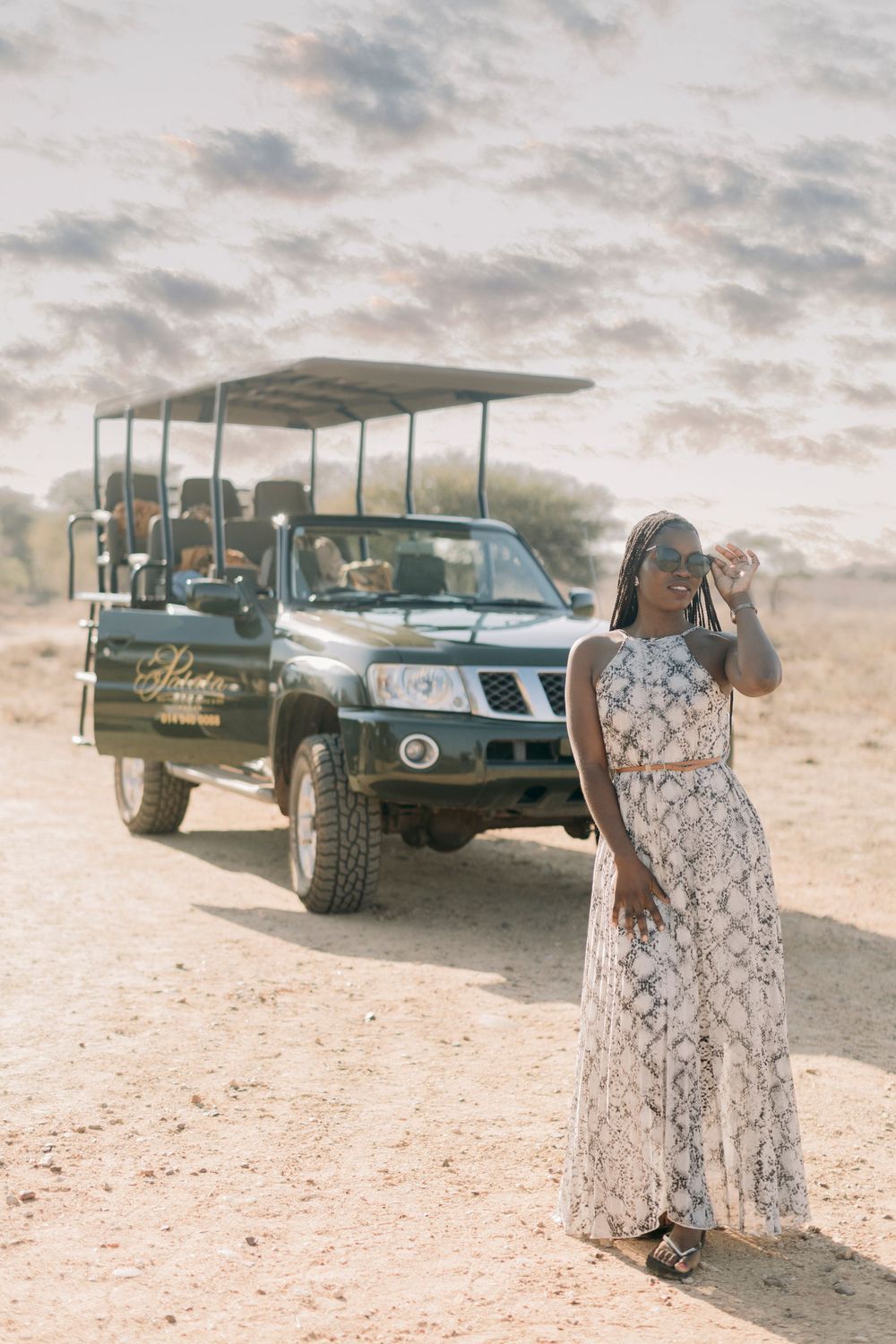 Stylish woman in white floral maxi dress posing in front of a safari game drive vehicle on dusty terrain.