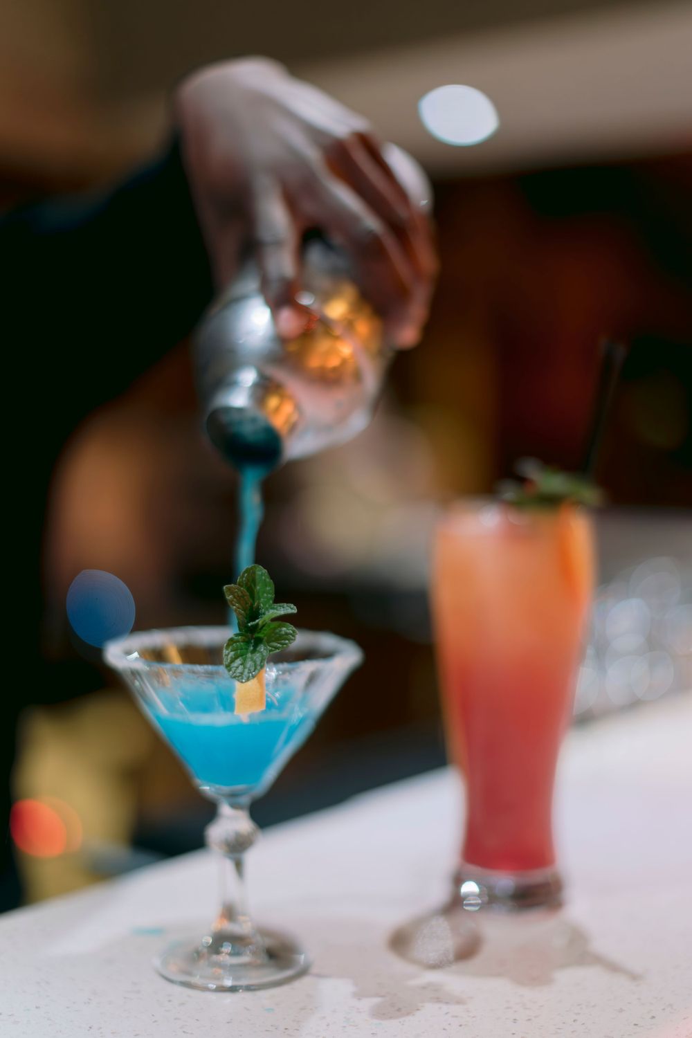 Bartender pouring a blue cocktail into a martini glass garnished with mint, beside a red drink.