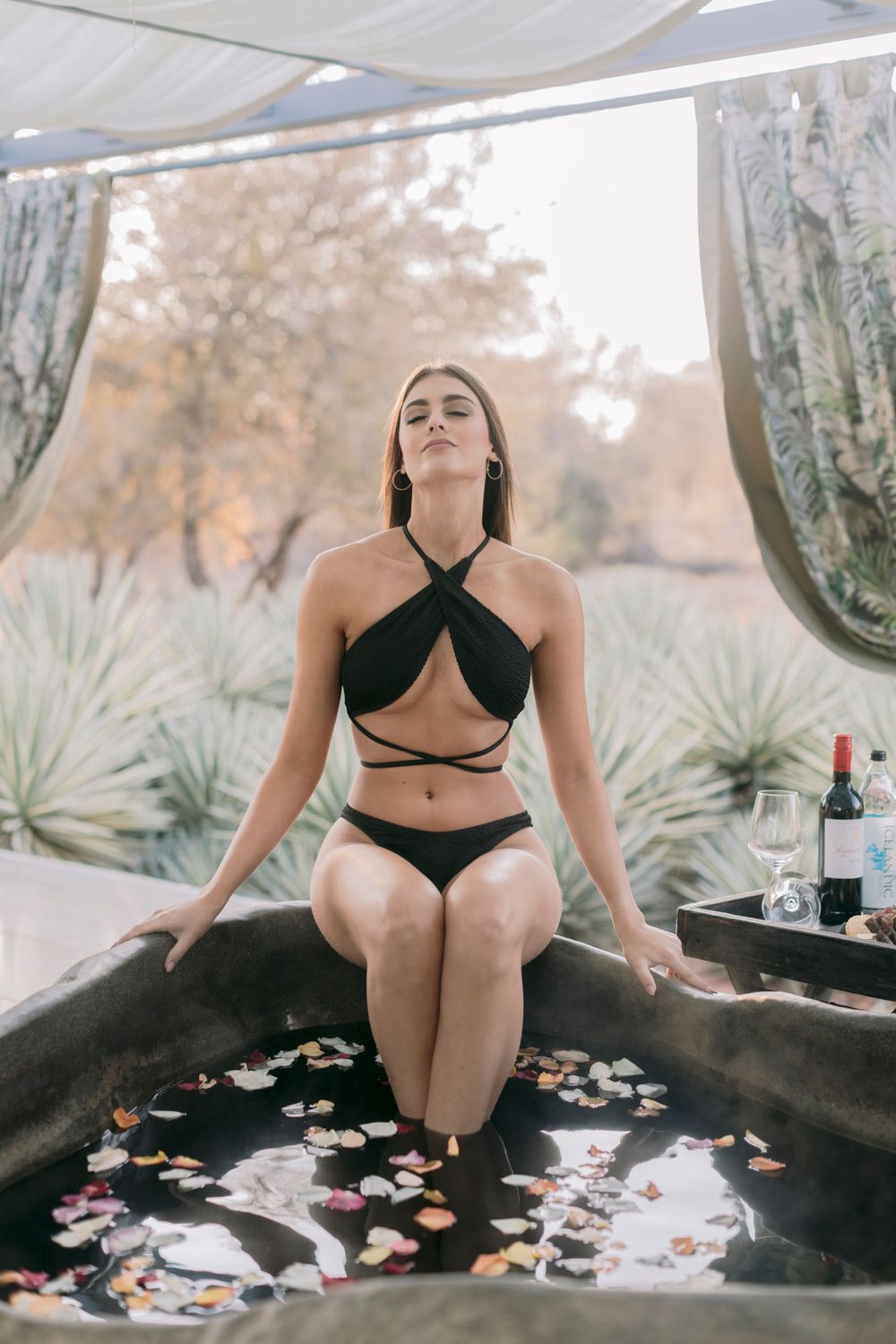 Woman in black bikini sitting in a floral petal bath with wine bottle nearby outdoors.