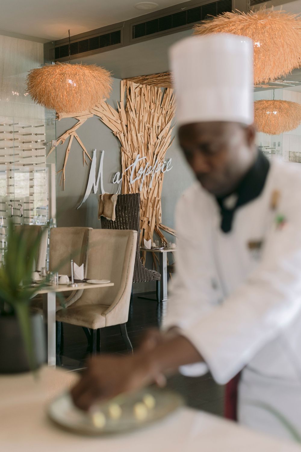 Chef in white uniform and toque plating a dish at an elegant fine dining restaurant interior.