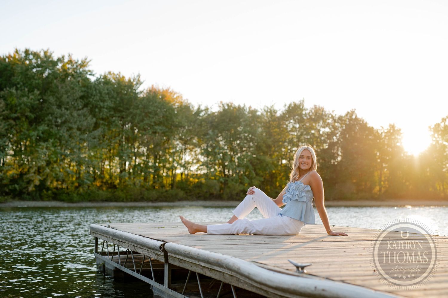 Person in summer outfit lounging on wooden dock with lake and sunset in background.