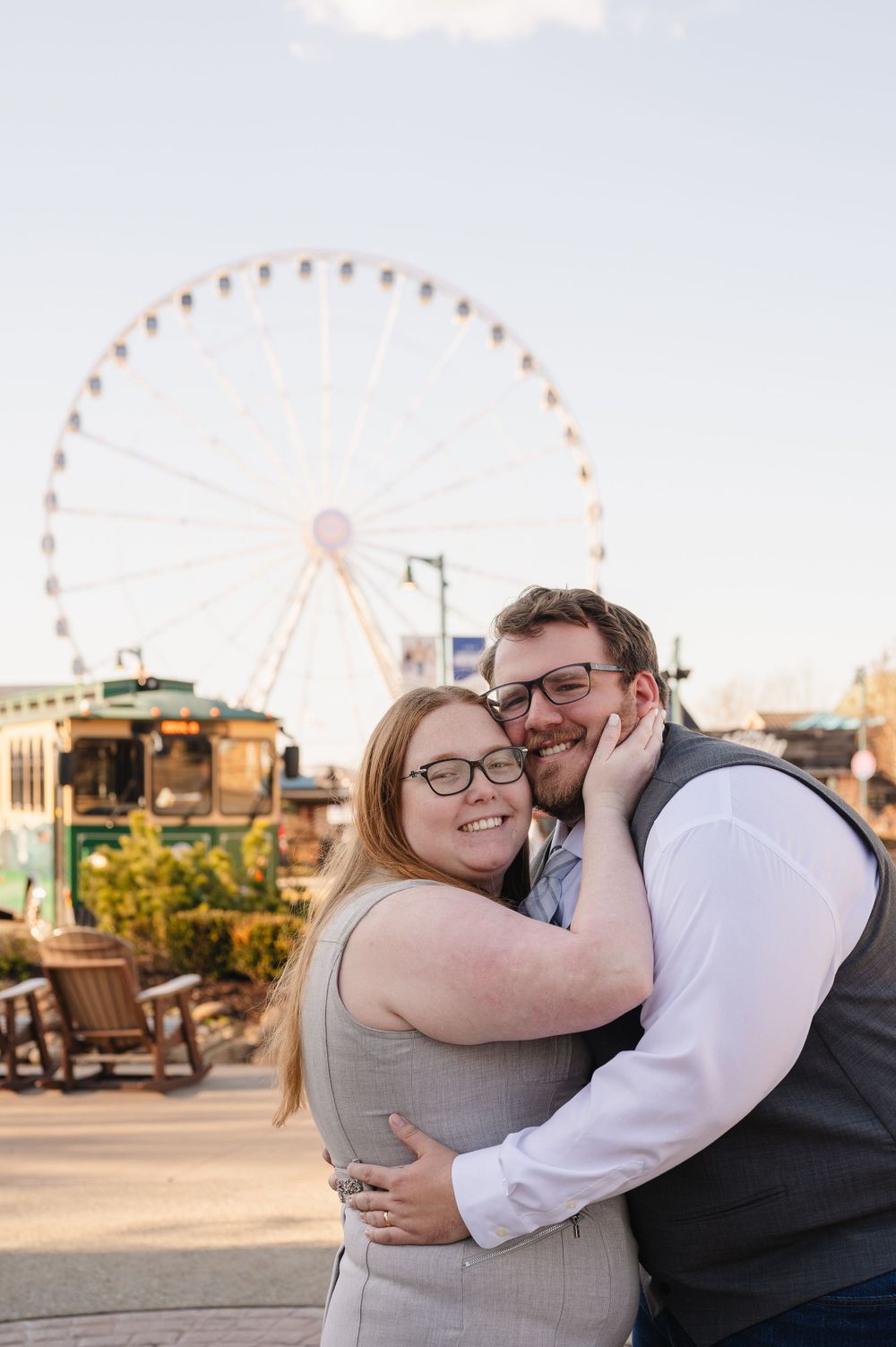 A couple embraces and smiles in front of a ferris wheel at a fairground during golden hour.