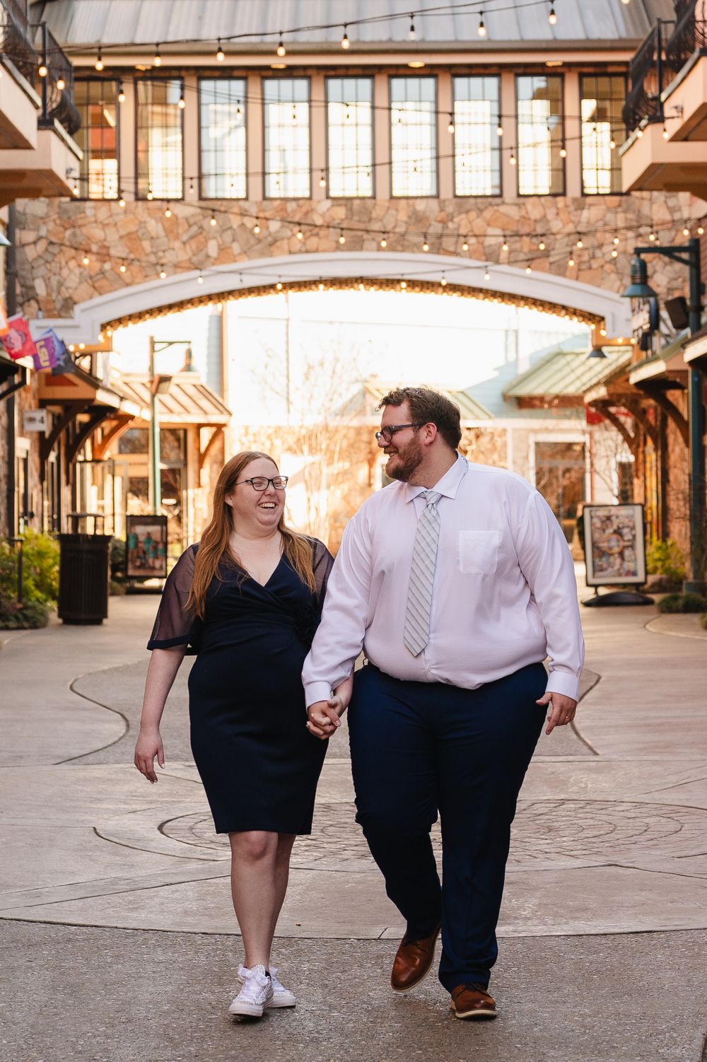 A couple walks hand in hand through a stylish shopping center with stone archways and warm lighting.