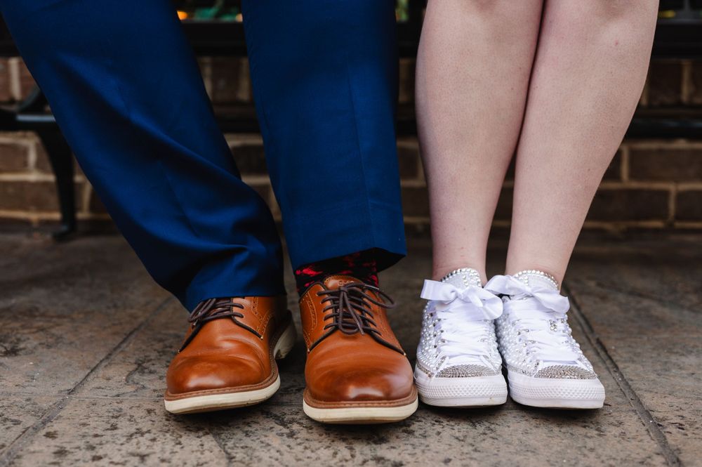 Two pairs of contrasting feet wearing brown leather dress shoes and white canvas sneakers side by side.