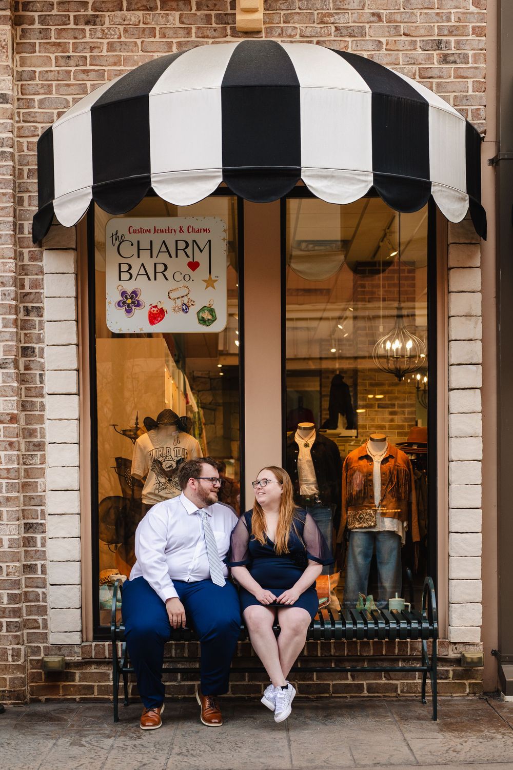 A small retail storefront with a black and white striped awning and 'Charm Bar' sign displaying jewelry in the window.