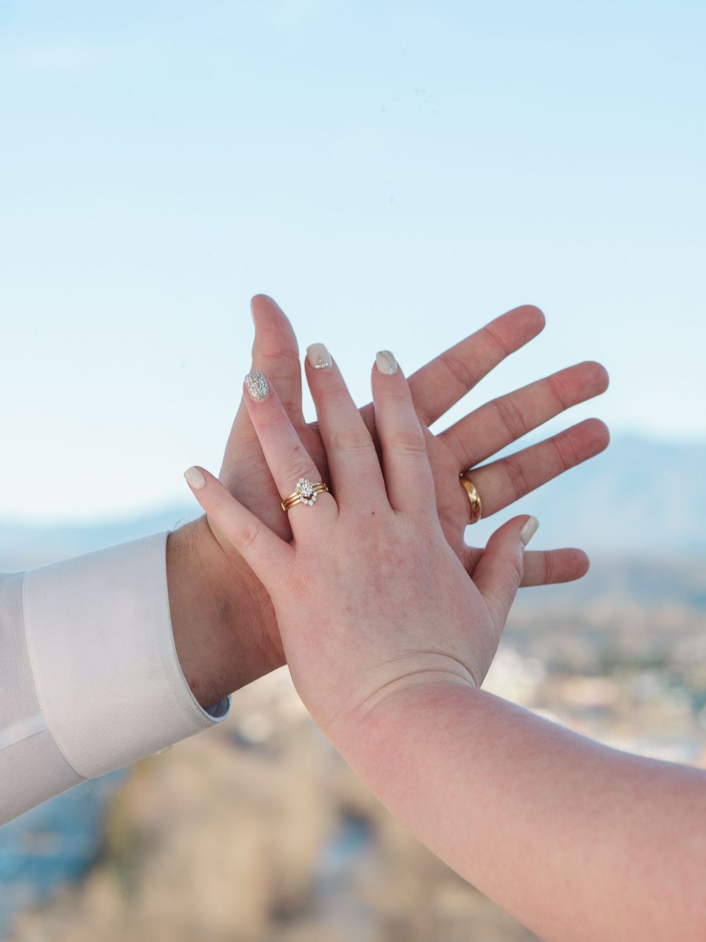 Two hands with elegant nail polish and rings touch tenderly against a blurred cityscape background.