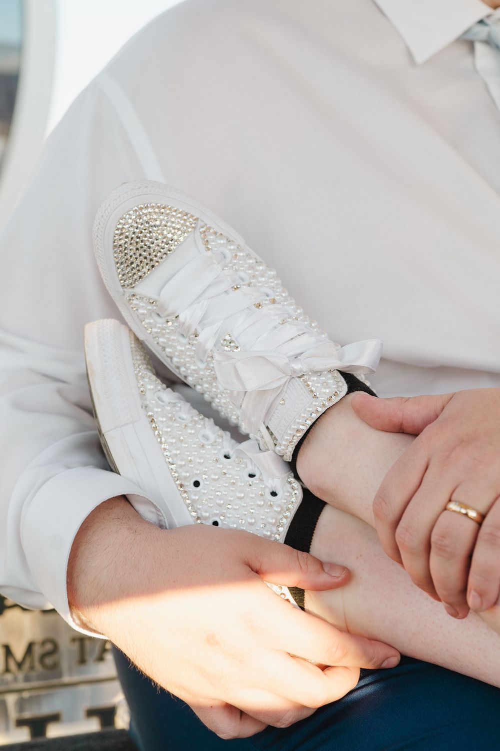 Close-up of hands tying laces on sparkly white sneakers decorated with pearls and crystals, with a gold wedding ring visible.