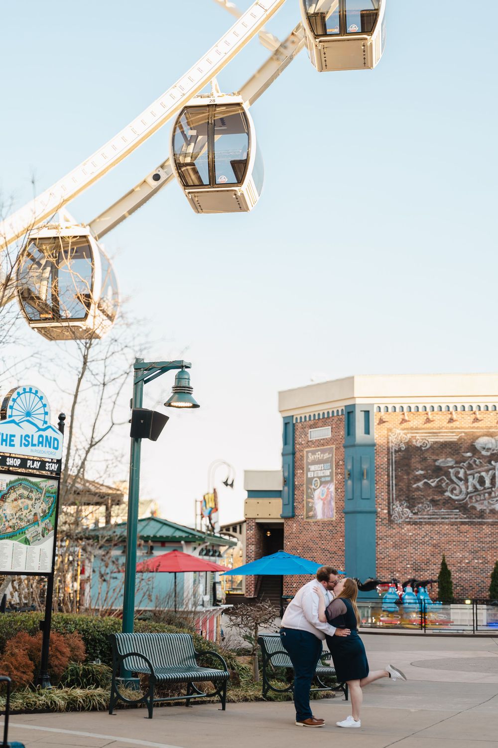 Couple embraces near a ferris wheel and lamp post in an urban plaza with brick buildings and benches.
