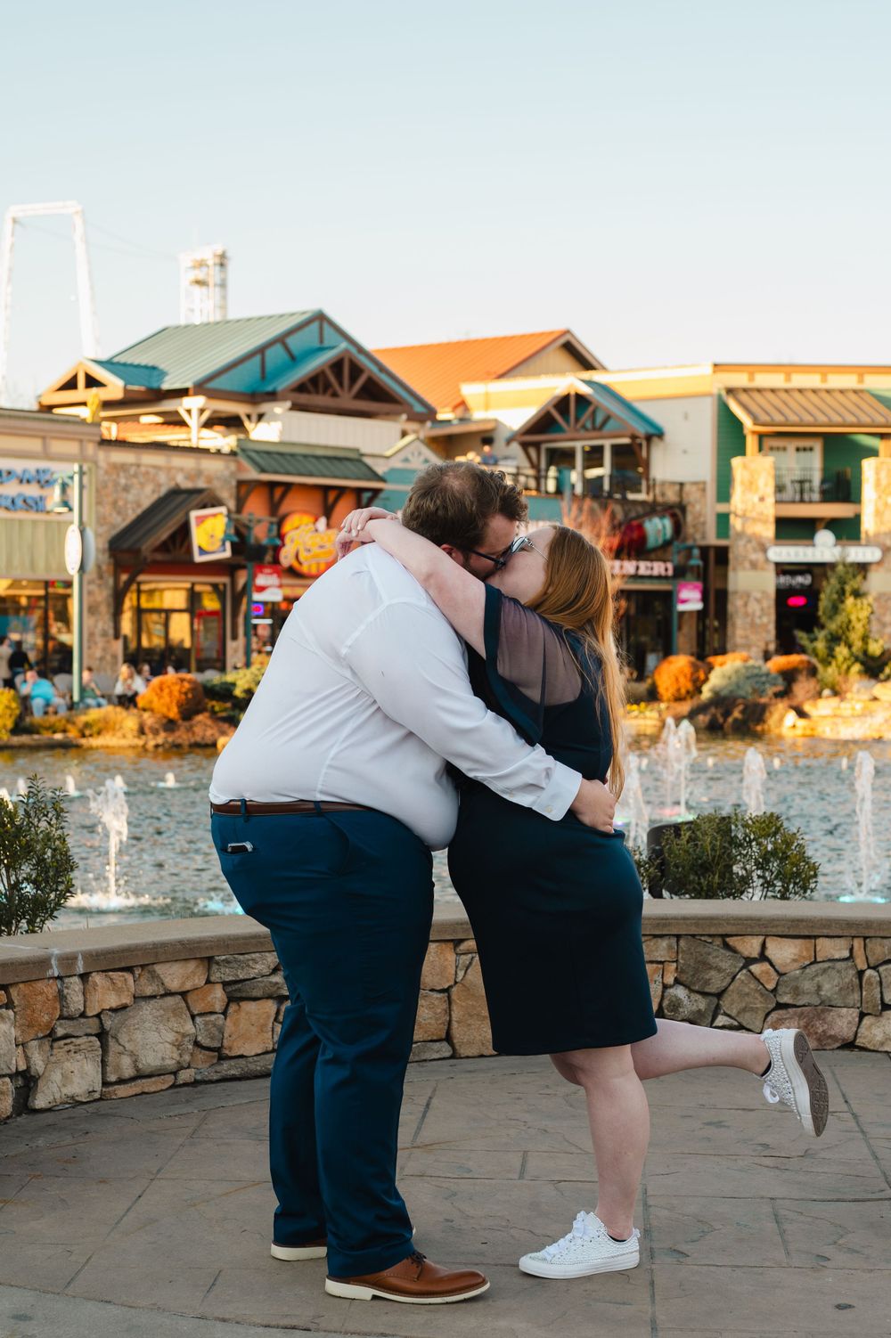 A couple embraces near a fountain at an outdoor shopping center with storefronts and pumpkin displays at sunset.