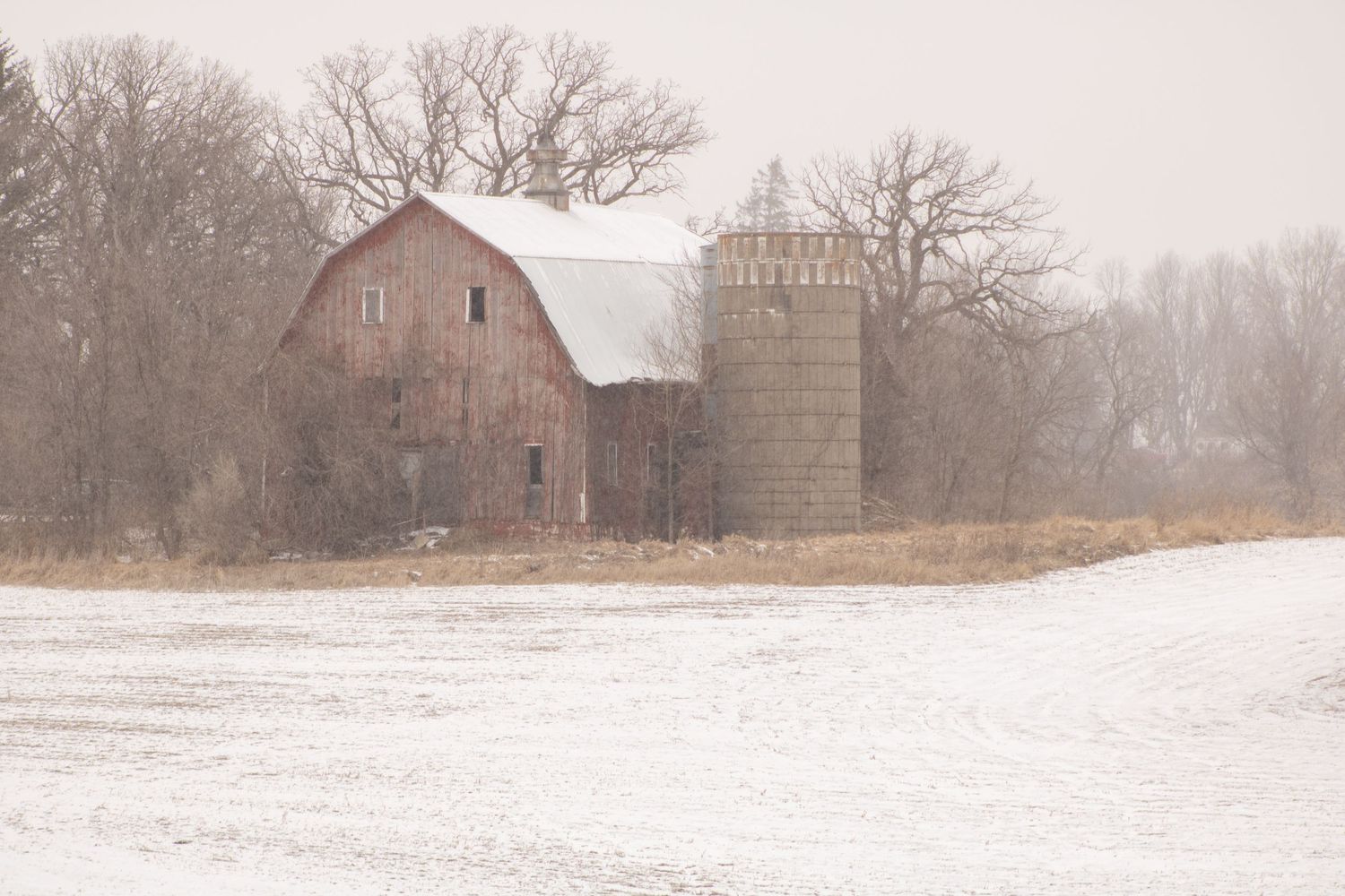 An old barn and silo stand in a snowy winter landscape.