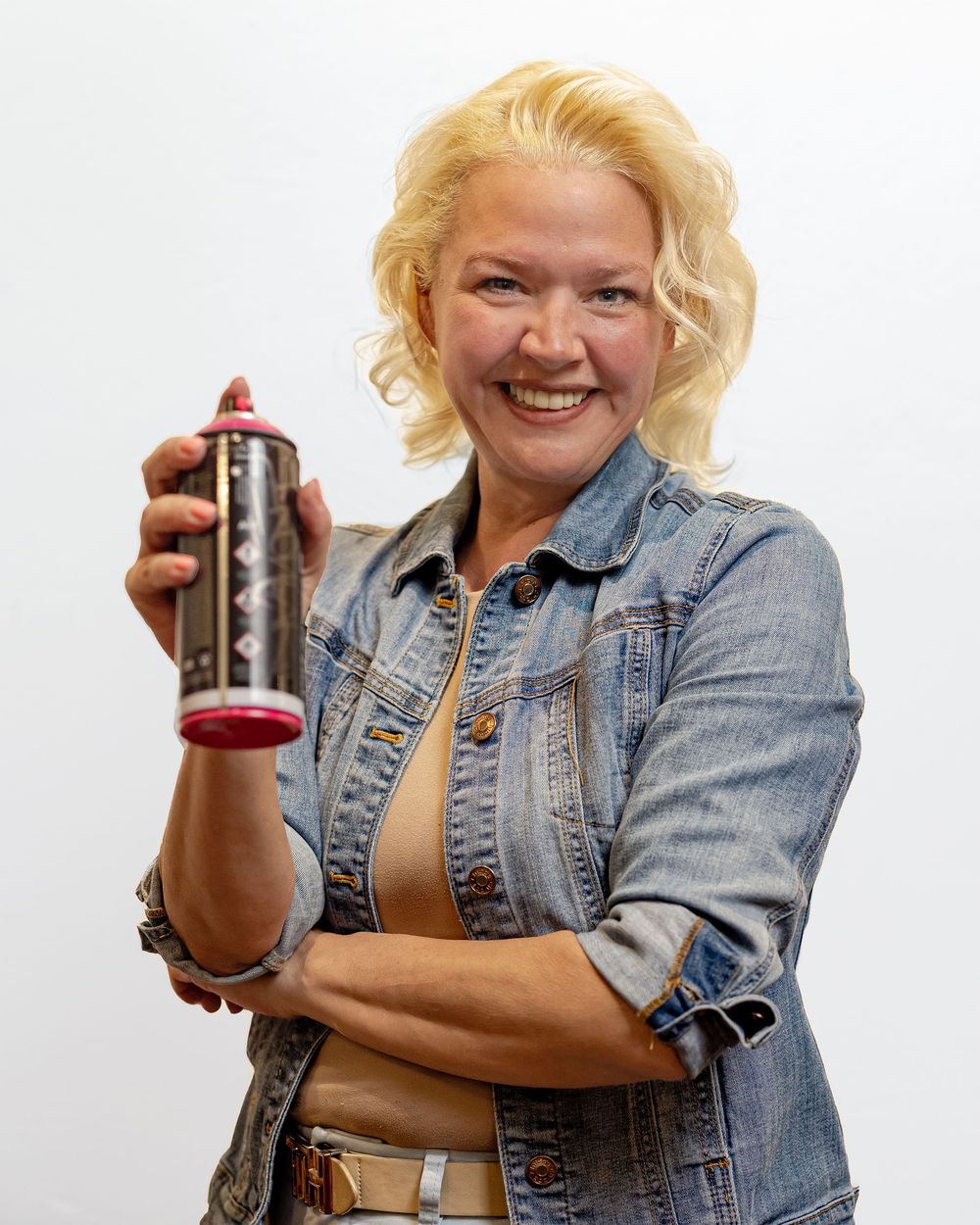 Smiling blonde woman in denim jacket holding a pink spray paint can against a white background.