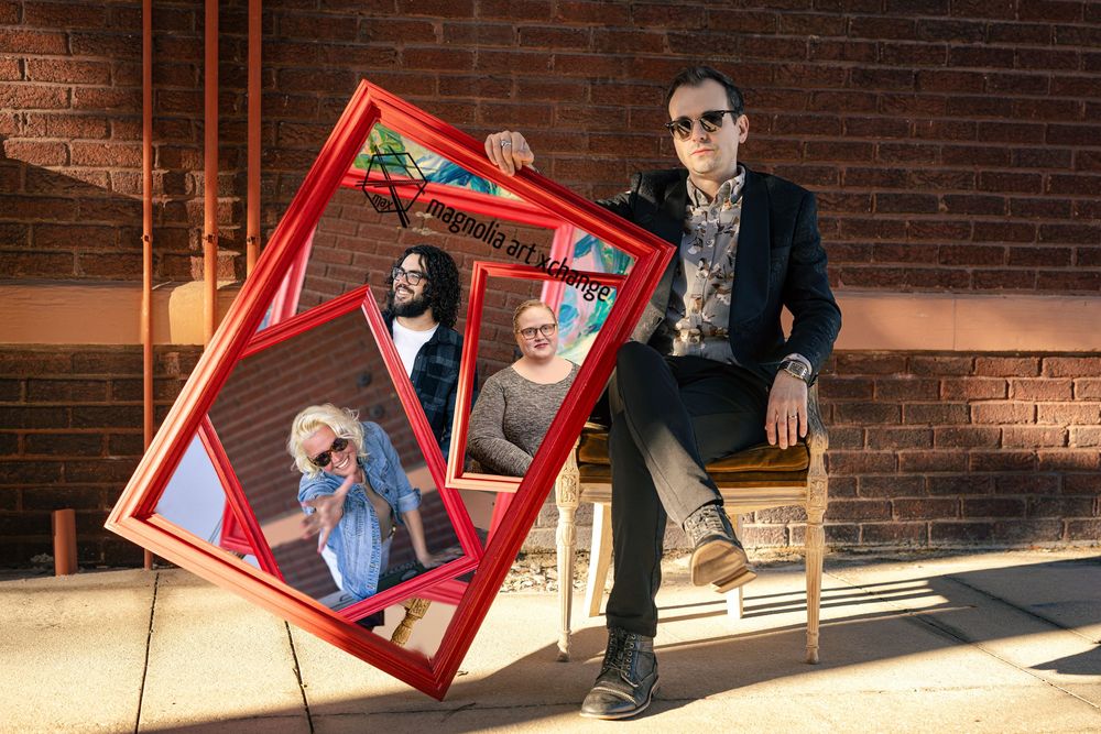 A man sits holding a large red picture frame containing reflections of people outdoors by a brick wall.