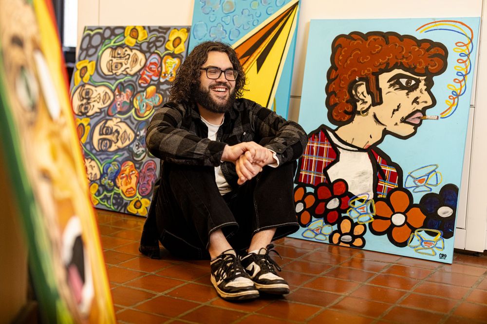 Artist smiling among colorful pop-art paintings including Bob Dylan portrait in bright studio.