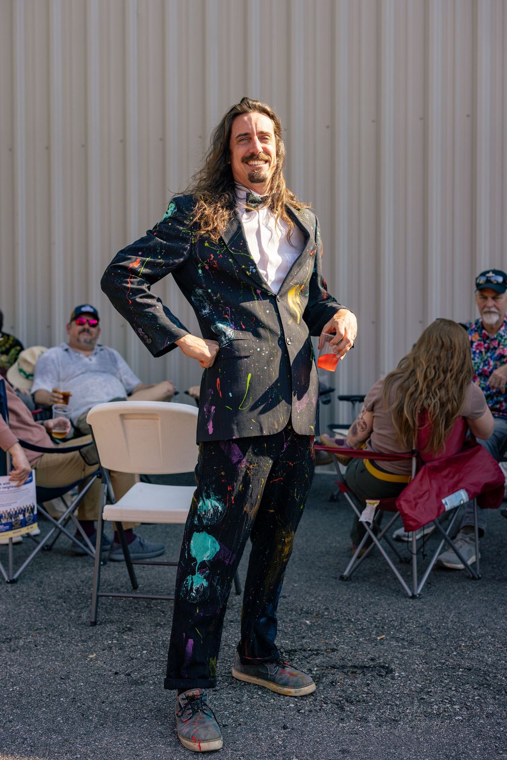 Man in paint-splattered black suit with long hair posing confidently at an outdoor art event.