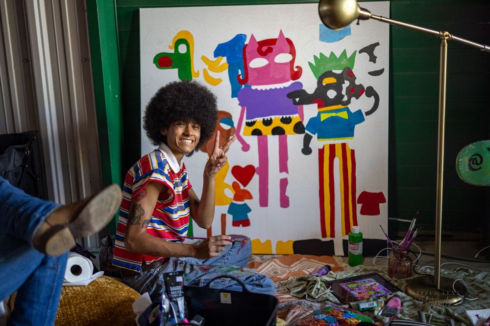 Artist smiling beside colorful monster painting in studio, surrounded by art supplies on floor.