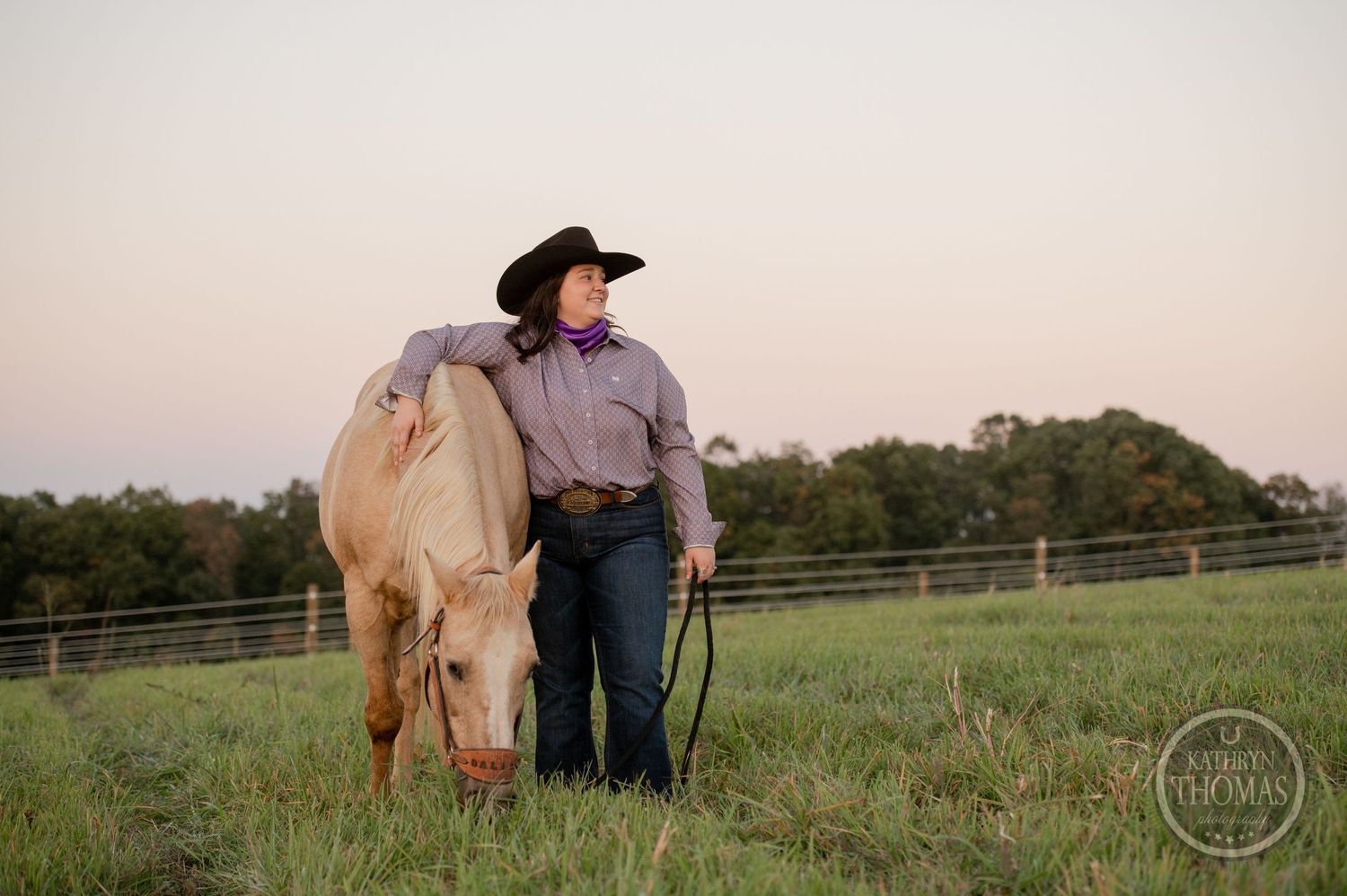 A person in western wear stands with a light-colored horse in a grassy fenced pasture at sunset.