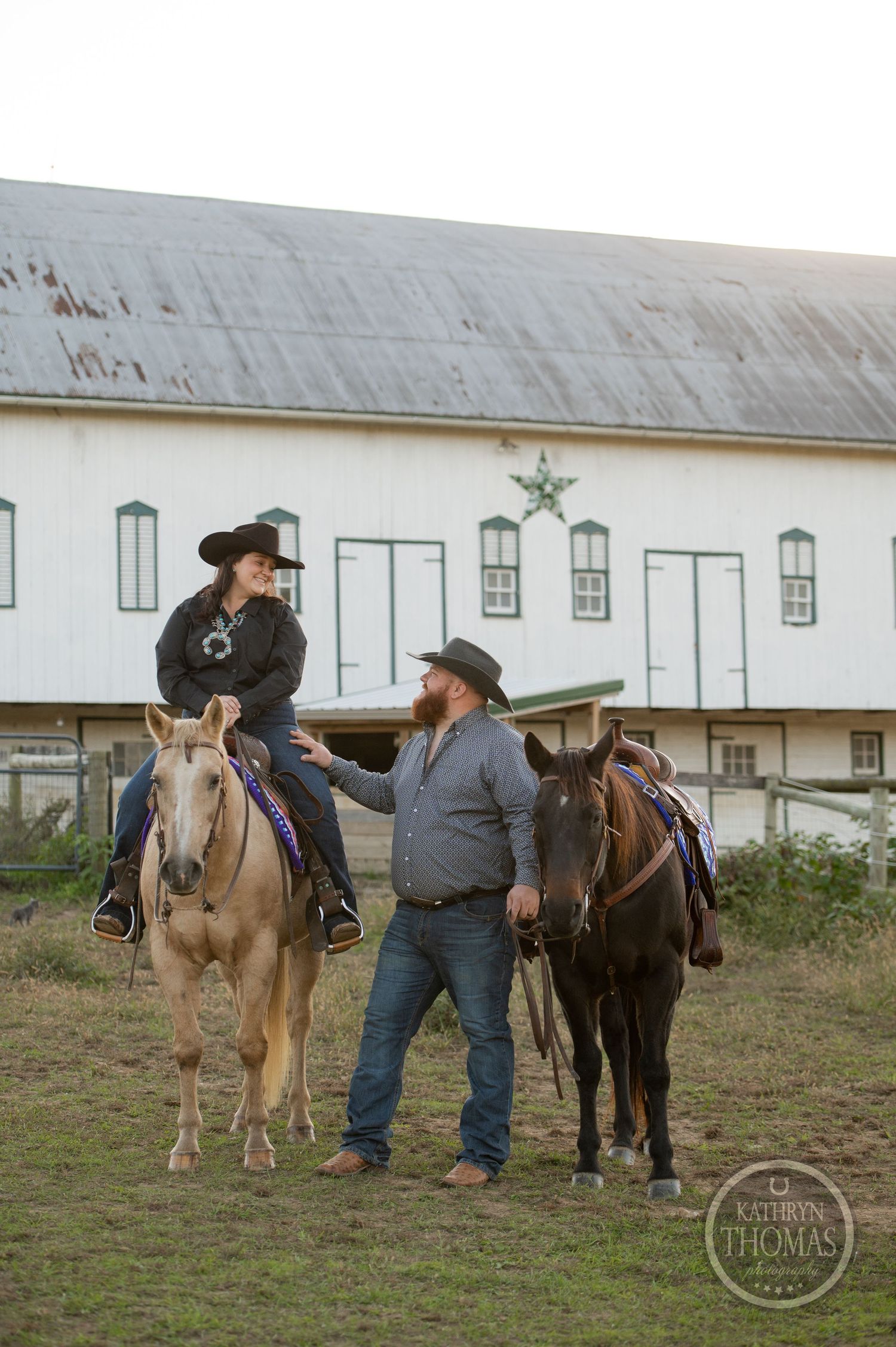 Mounted rider and companion with horses chat in front of large white barn.