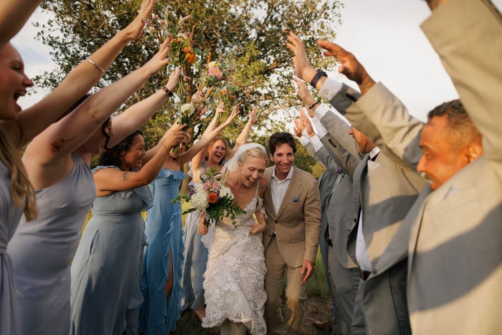 Joyful wedding party celebrates with raised arms as bride and groom walk through a festive outdoor arch.