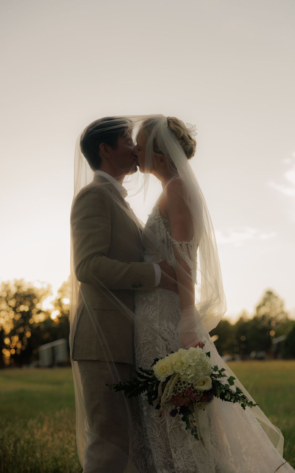 Bride and groom share a romantic kiss under a flowing veil at sunset in an open field.
