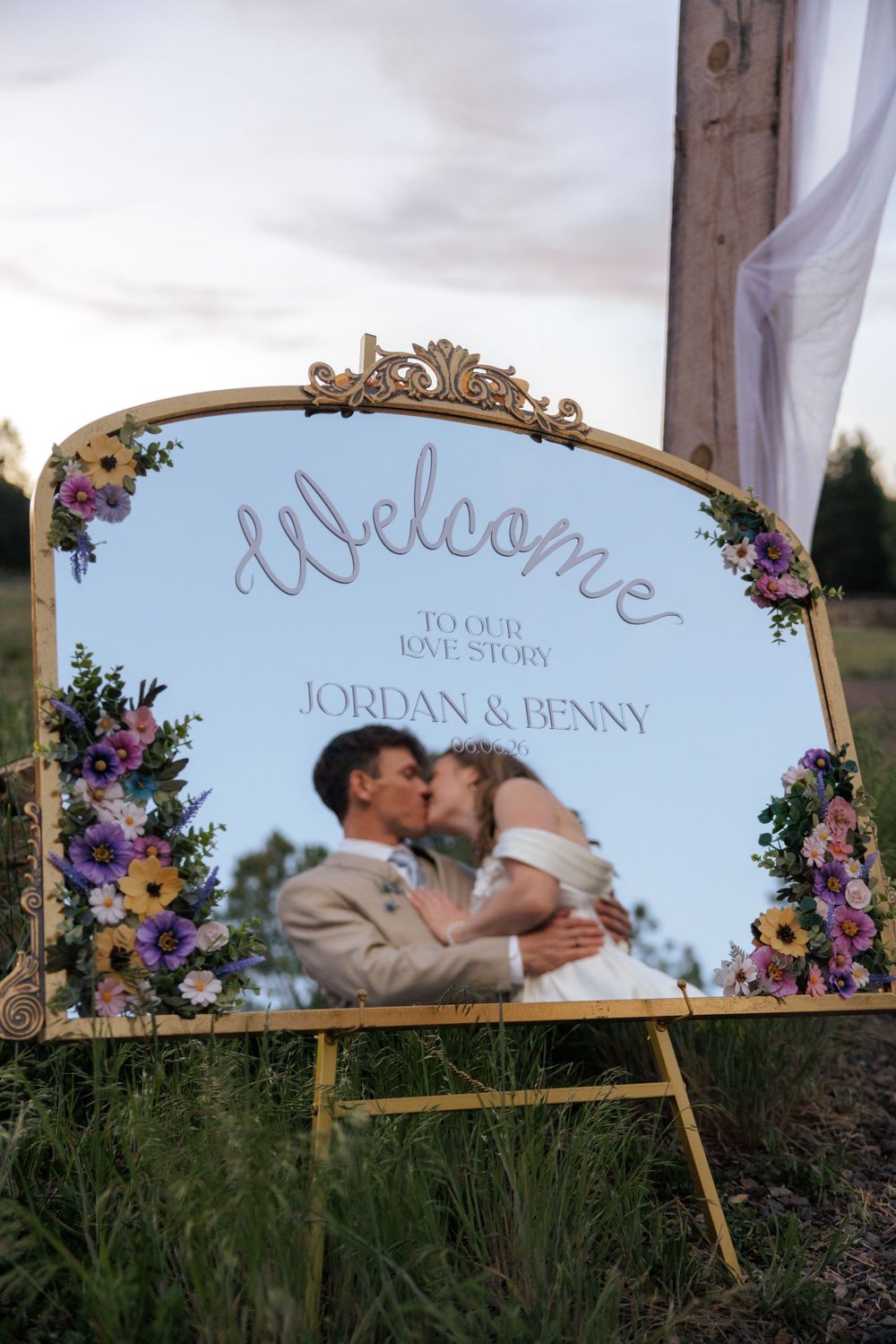 Bridge and Groom kiss, as seen through an elegant gold-framed welcome mirror sign decorated with flowers.