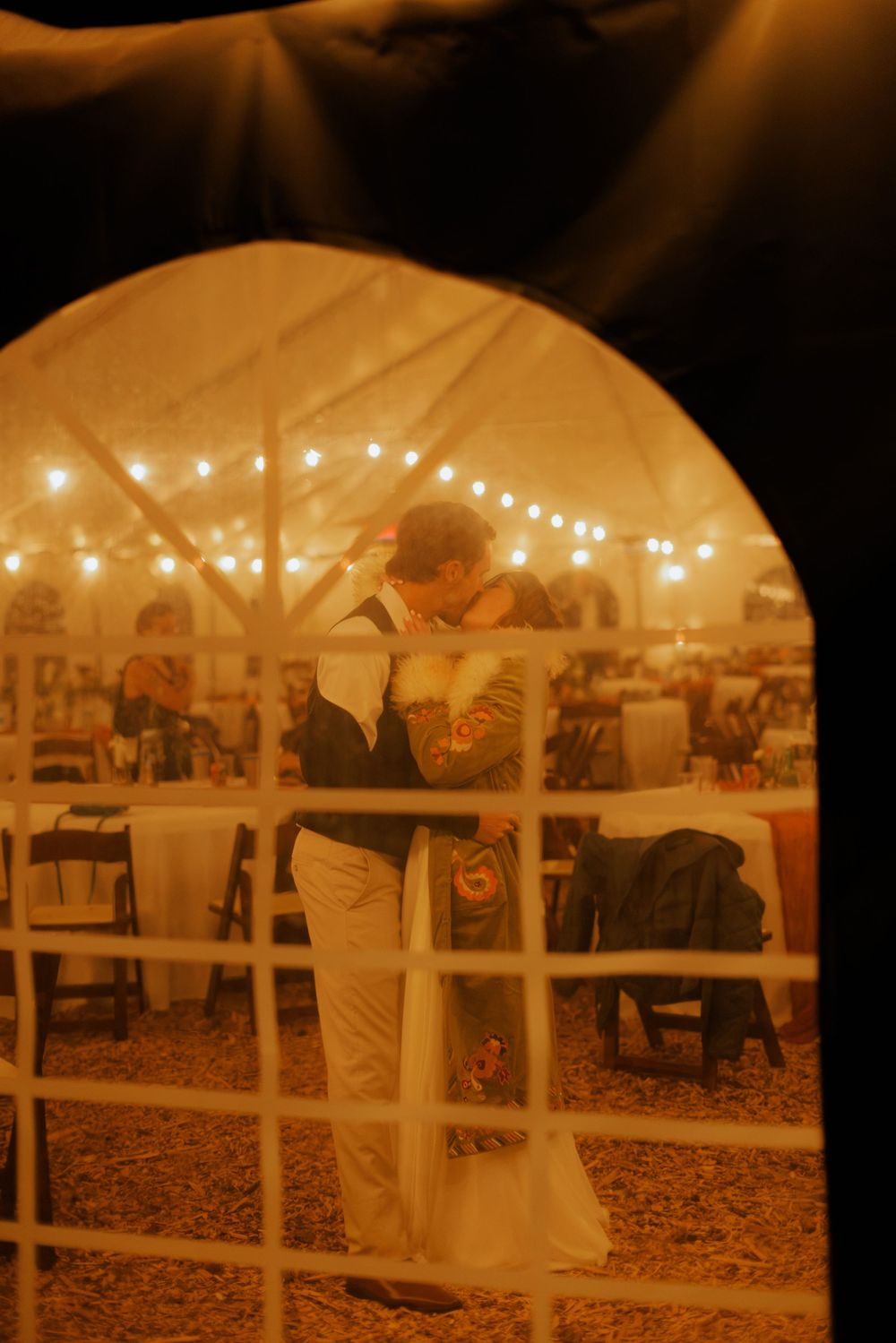Couple sharing first dance under string lights in a warmly lit wedding reception tent, viewed through an arched window.