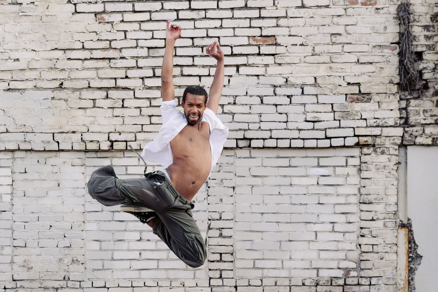 Shirtless dancer performs powerful jumping pose against weathered white brick wall background.