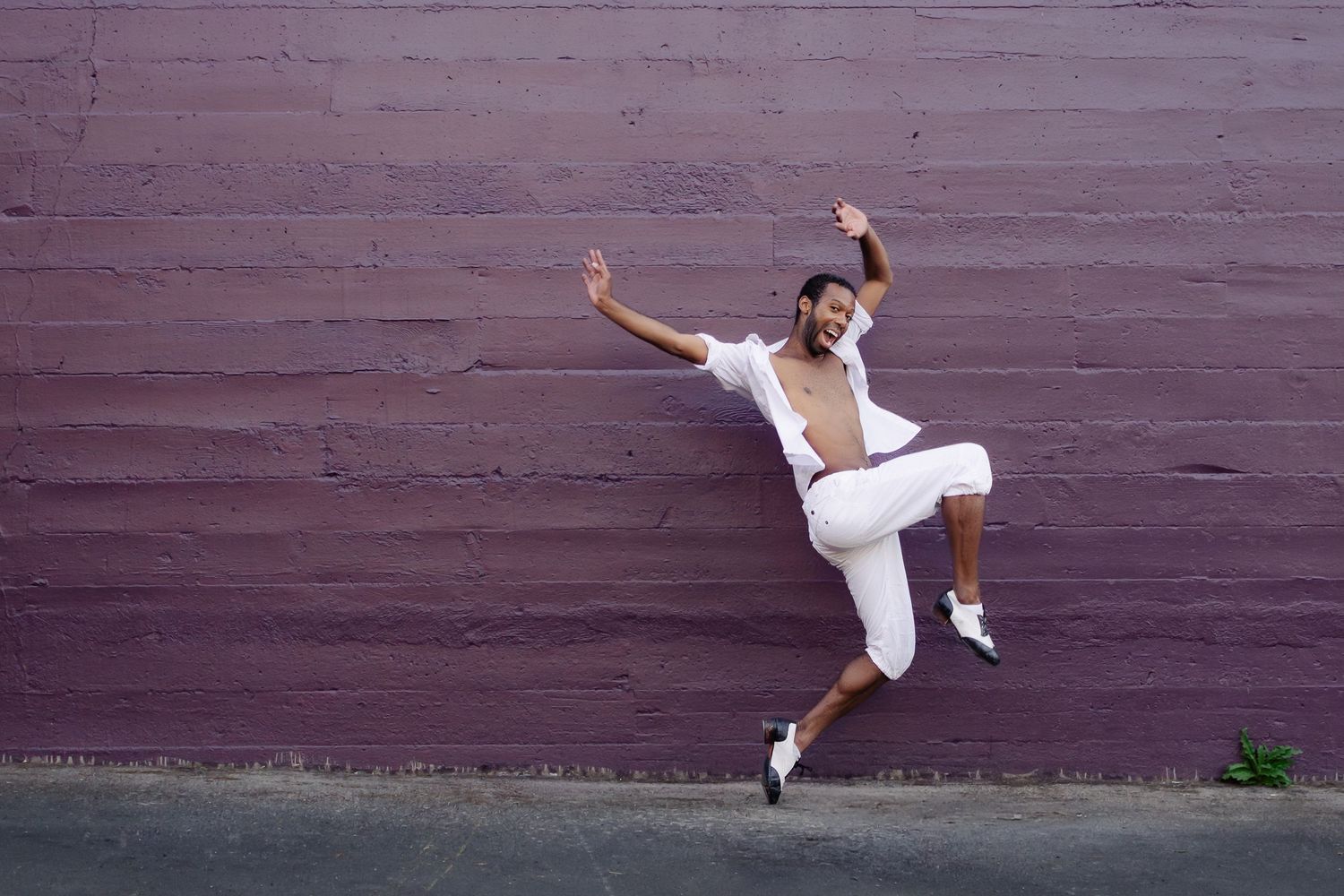 Energetic dancer in white outfit leaps gracefully against purple wall during urban dance performance.