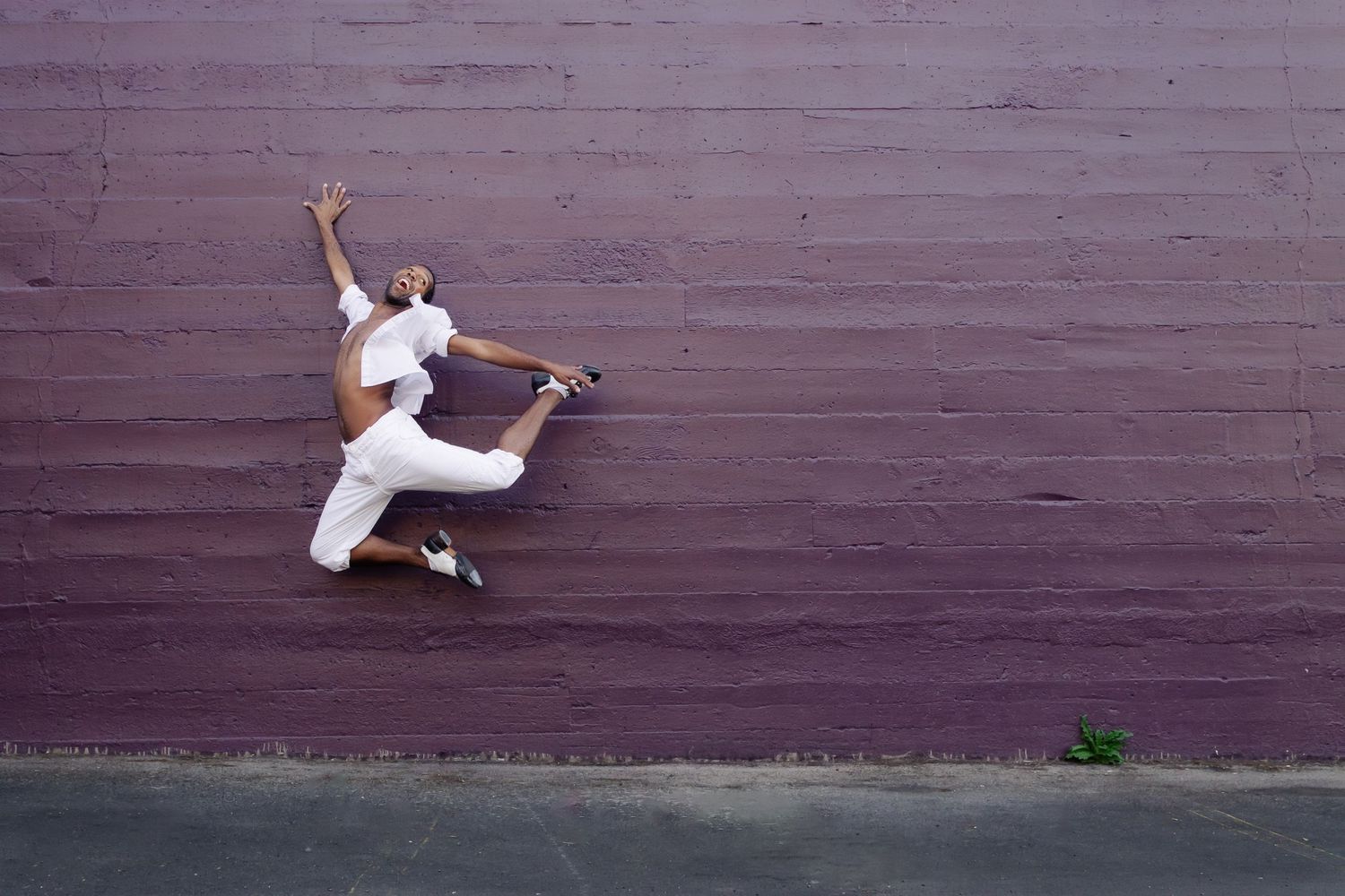 Dancer in white clothing leaps gracefully against a purple wall in an artistic urban dance pose.