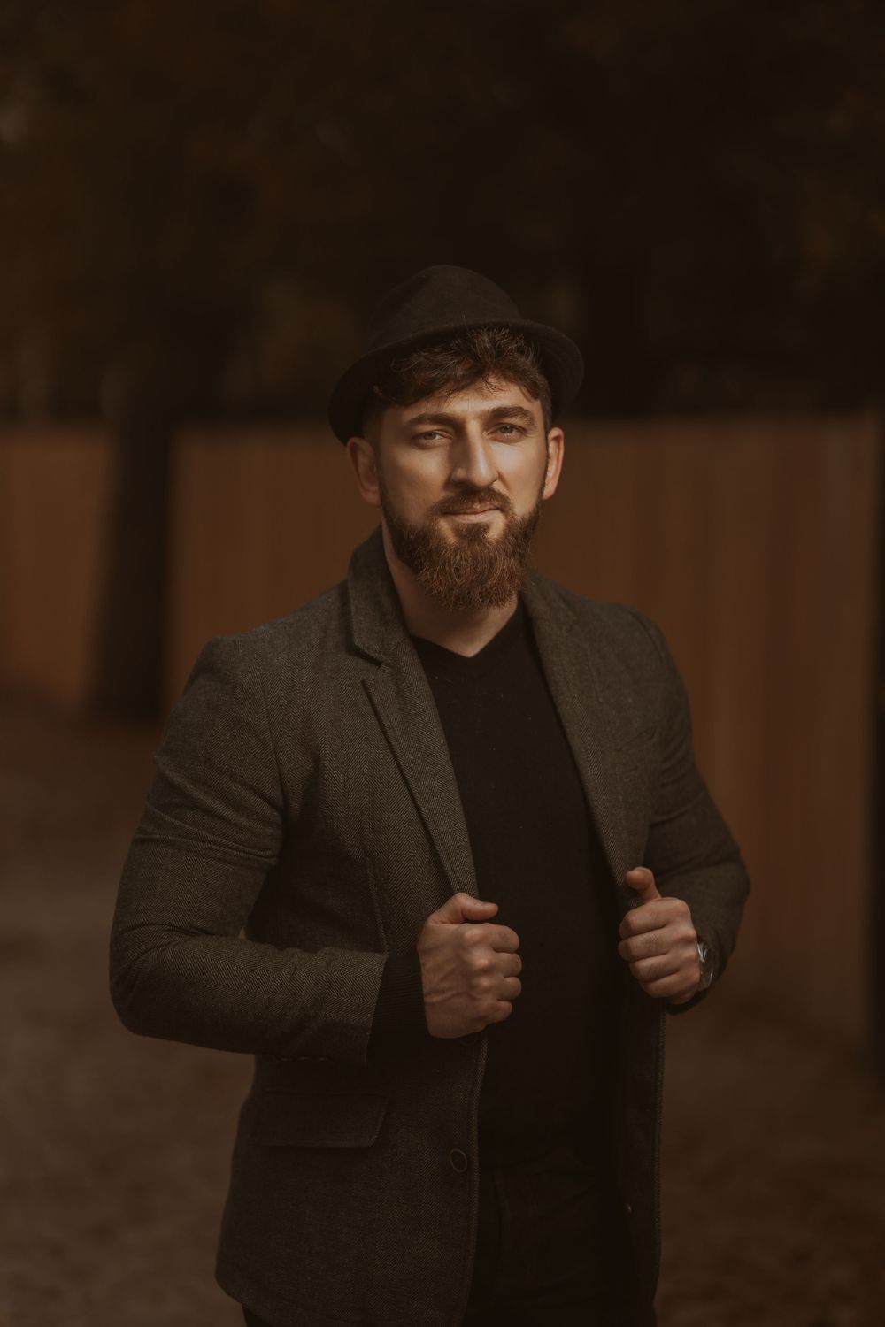 Portrait of bearded man in dark blazer and newsboy cap standing confidently against rustic backdrop.