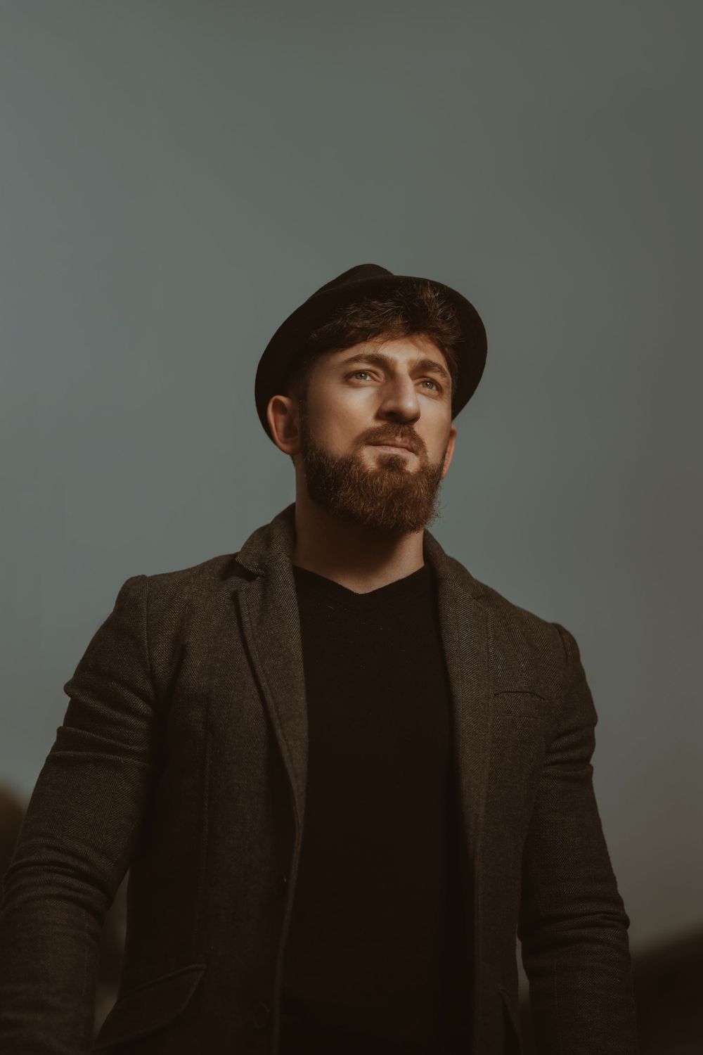 Professional portrait of someone in a dark coat and beanie looking upward against a grey background.
