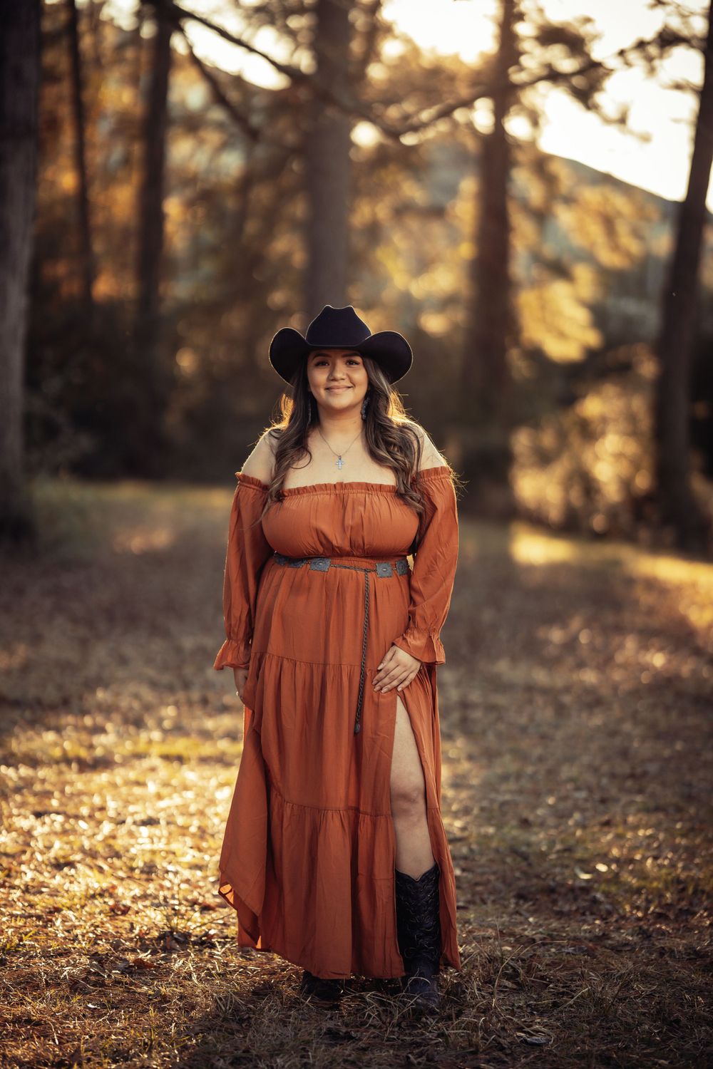 A person in a brown flowing maxi dress and black cowboy hat poses in a sun-dappled autumn forest.