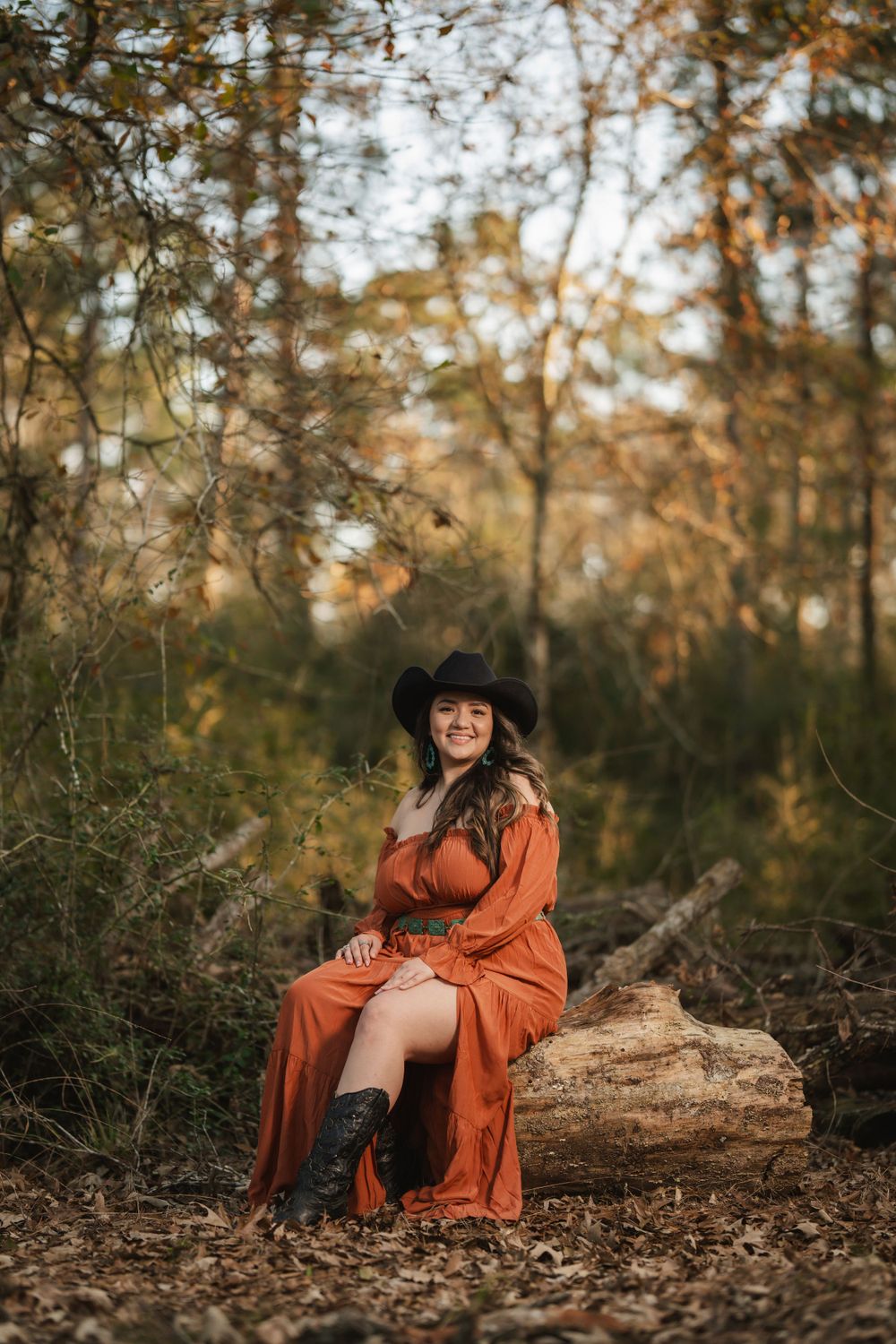 Someone wearing an orange dress and black hat sits on a fallen log in an autumn forest setting at golden hour.