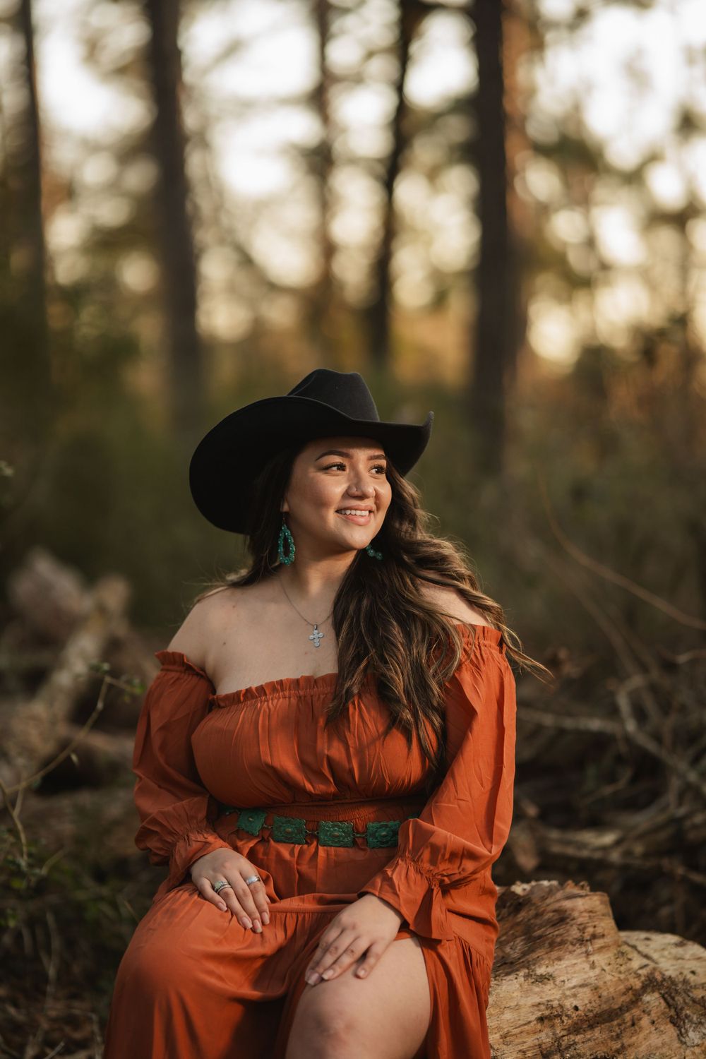 Woman in orange dress and black cowboy hat sits on log in forest during golden hour sunset.